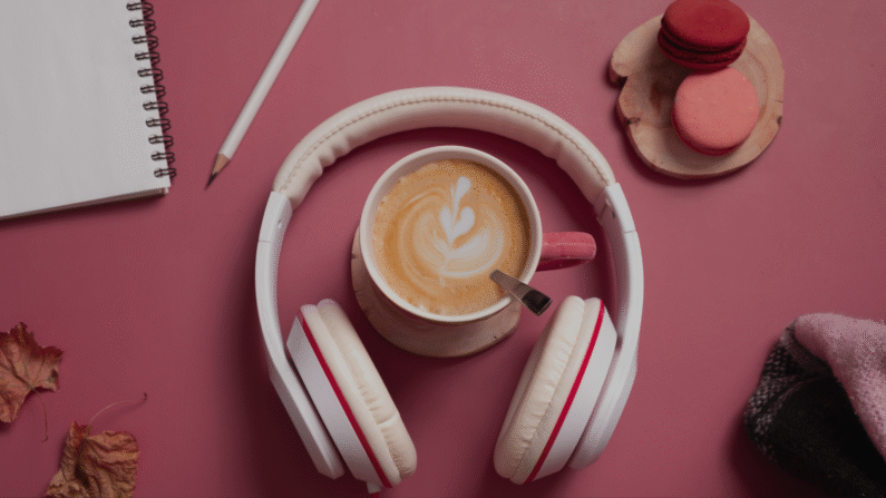 kawa cappuccino w różonwej filiżance, obok słuchawki w białym kolorze i różowe ciasteczka makaroniki, wszystko leży na różowym blacie