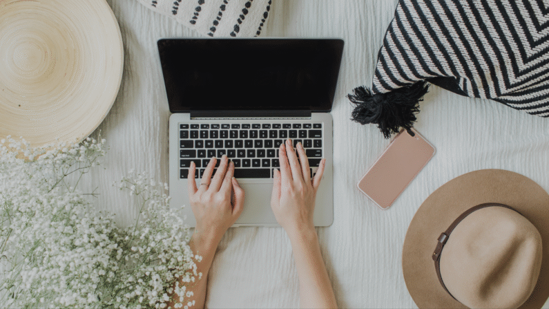 jasny koc, na nim otwarty laptop, dłonie na klawiaturze, dookoła rozłożone poduszki dekoracyjne, kapelusz, smartfon